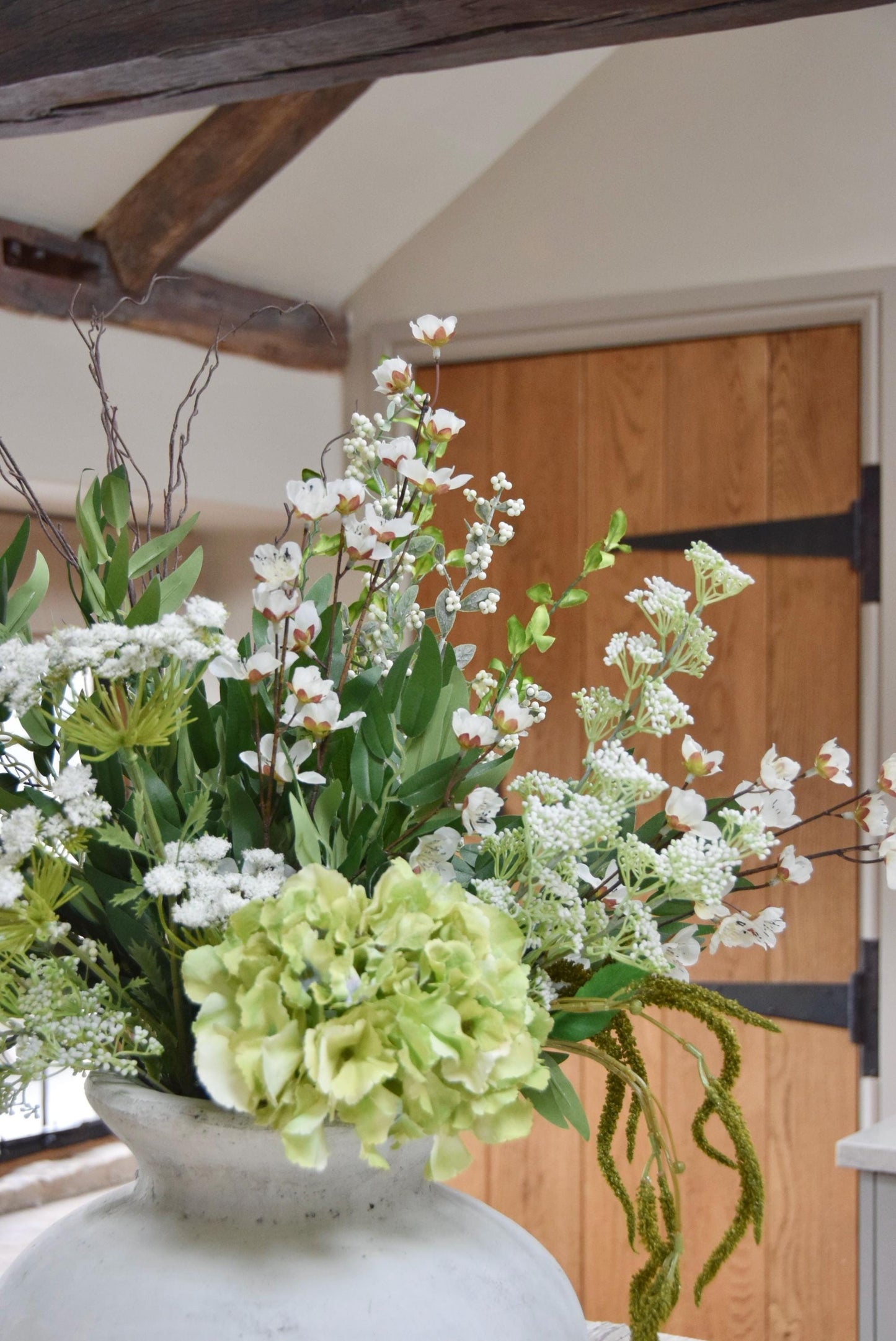 Stone antique vase placed on a kitchen countertop with an arrangement of green and white mixed flowers with lots of foliage. Displayed in a country style kitchen for home decor.