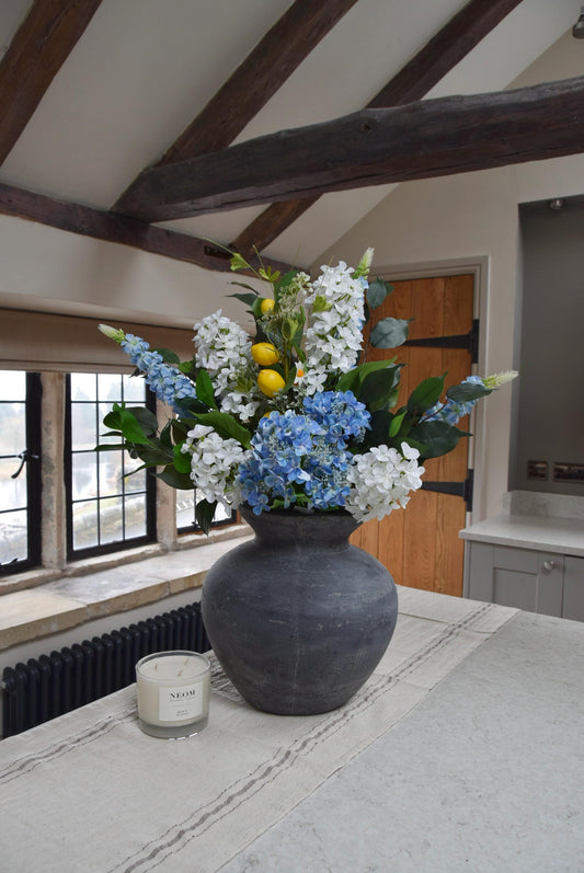 Grey antique vase placed on a kitchen countertop with arrangement of blue and white hydrangeas with lemons and green foliage. Displayed in a country style kitchen for home decor.