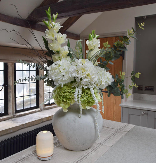 Stone antique vase placed on a kitchen countertop with an arrangement of white hydrangeas, green hydrangeas, cream gladioli and foliage. Displayed in a country style kitchen for home decor.