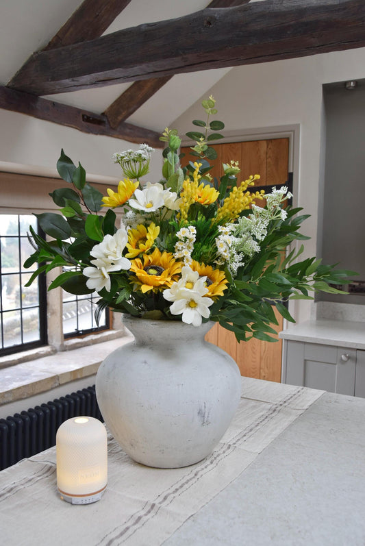 Stone antique vase placed on a kitchen countertop with an arrangement of yellow sunflowers, white wildflowers and green foliage. Displayed in a country style kitchen for home decor.