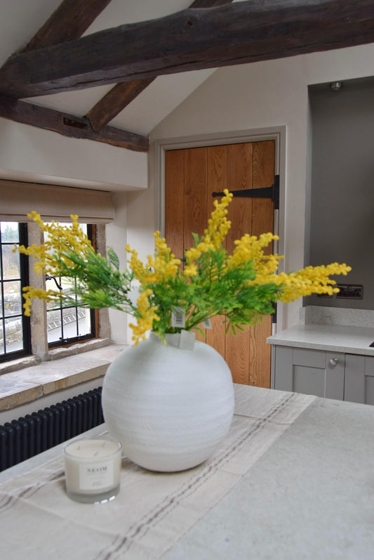Round white vase placed on a kitchen countertop with a bunch of yellow mimosa stems. Displayed in a country style kitchen for home decor.
