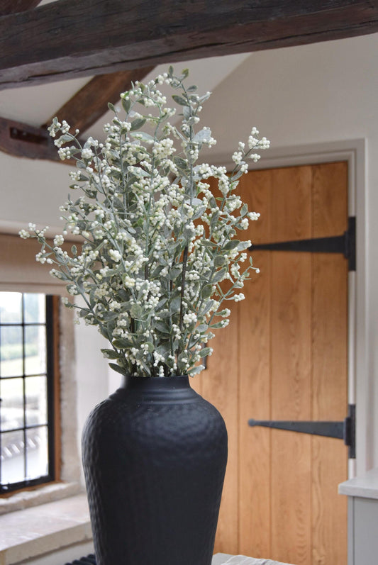 Tall black vase placed on a kitchen countertop with a bunch of artificial berry leaf stems. Displayed in a country style kitchen for home decor.
