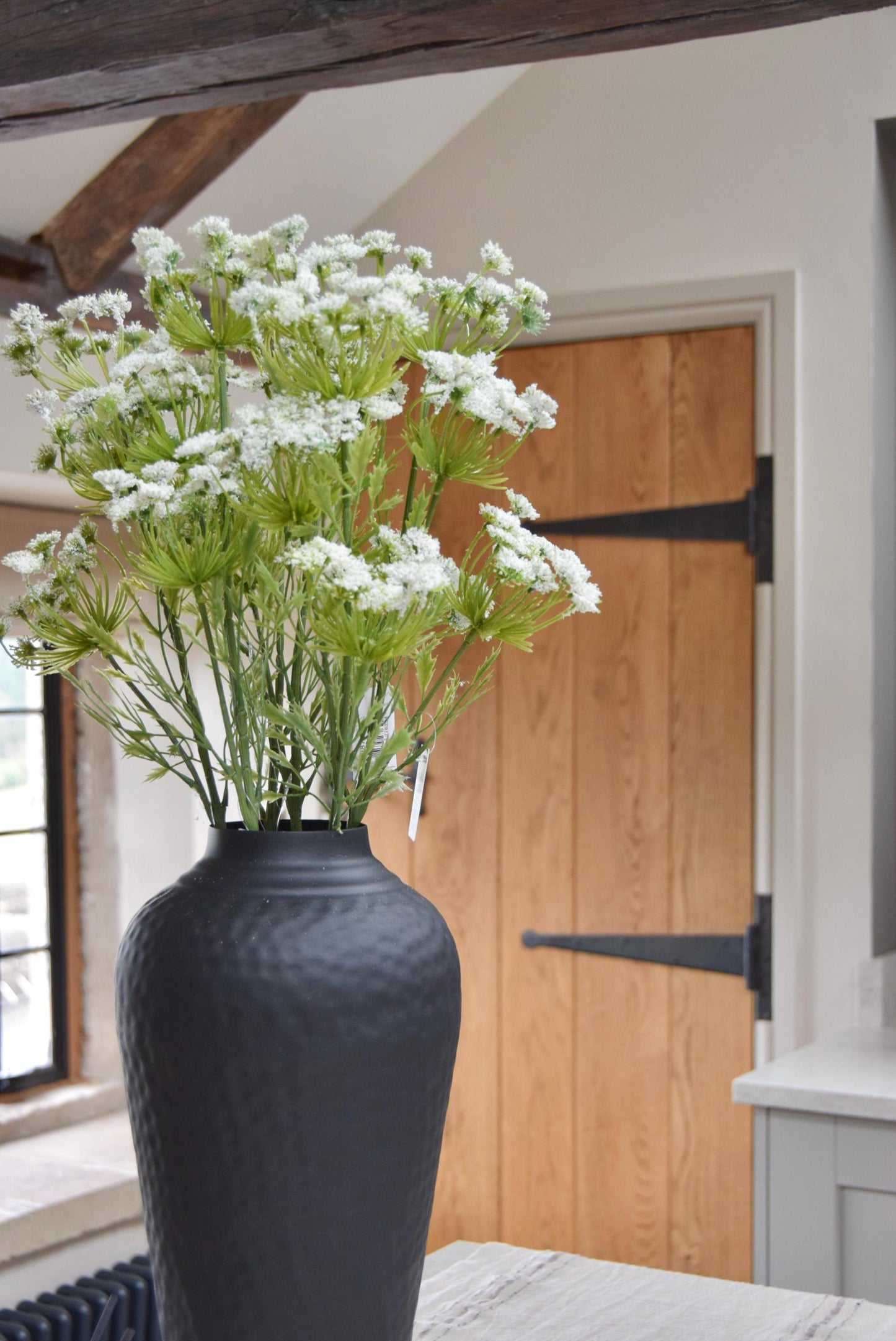 Tall black vase placed on a kitchen countertop with a bunch of white cow parsley. Displayed in a country style kitchen for home decor.