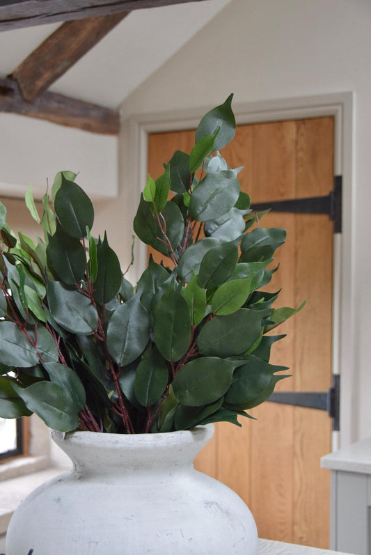 Stone antique vase placed on a kitchen countertop with a bunch of artificial bay leaf sprays. Displayed in a country style kitchen for home decor.