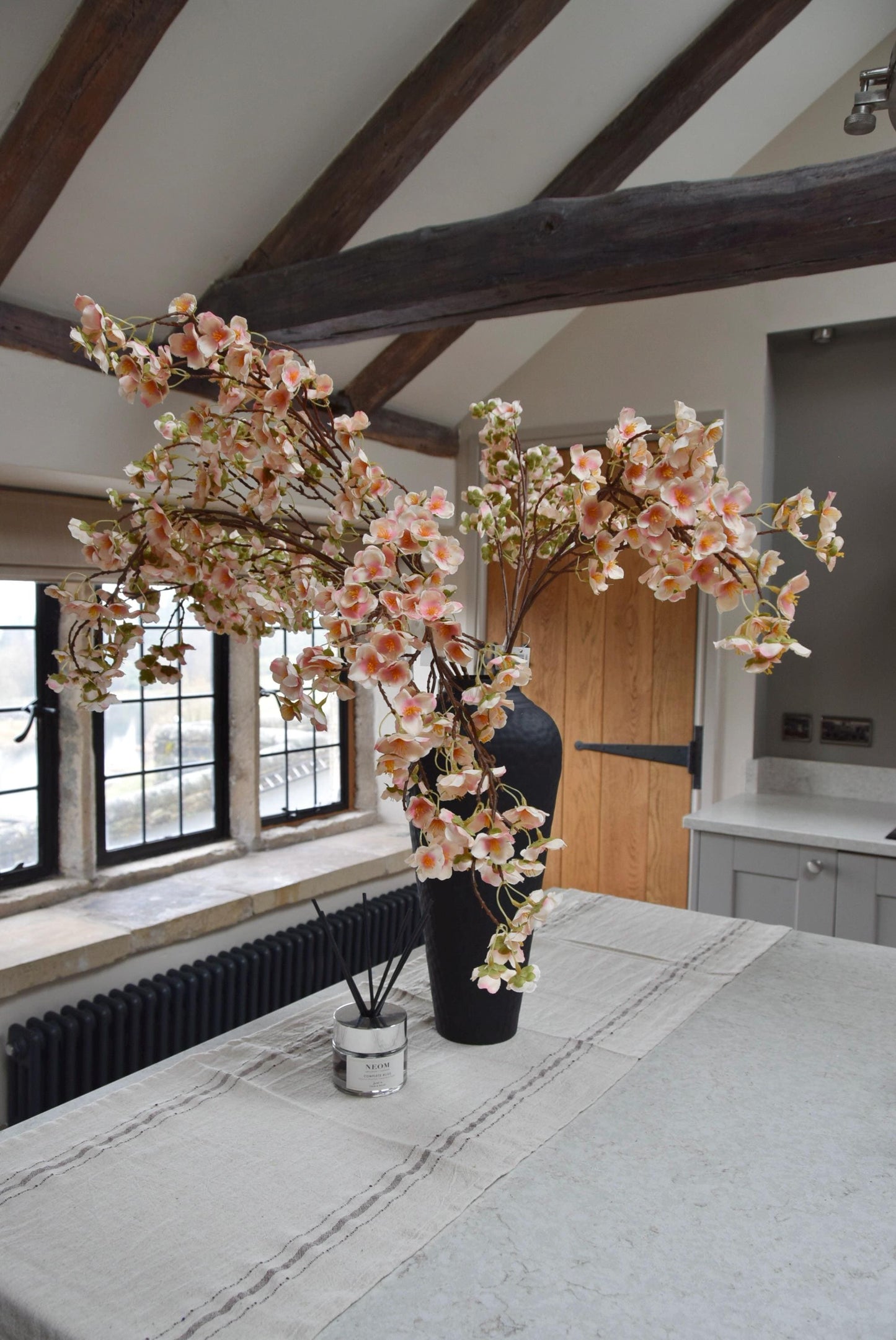 Tall black vase placed on a kitchen countertop with a bunch of hanging pink plum blossom stems. Displayed in a country style kitchen for home decor.