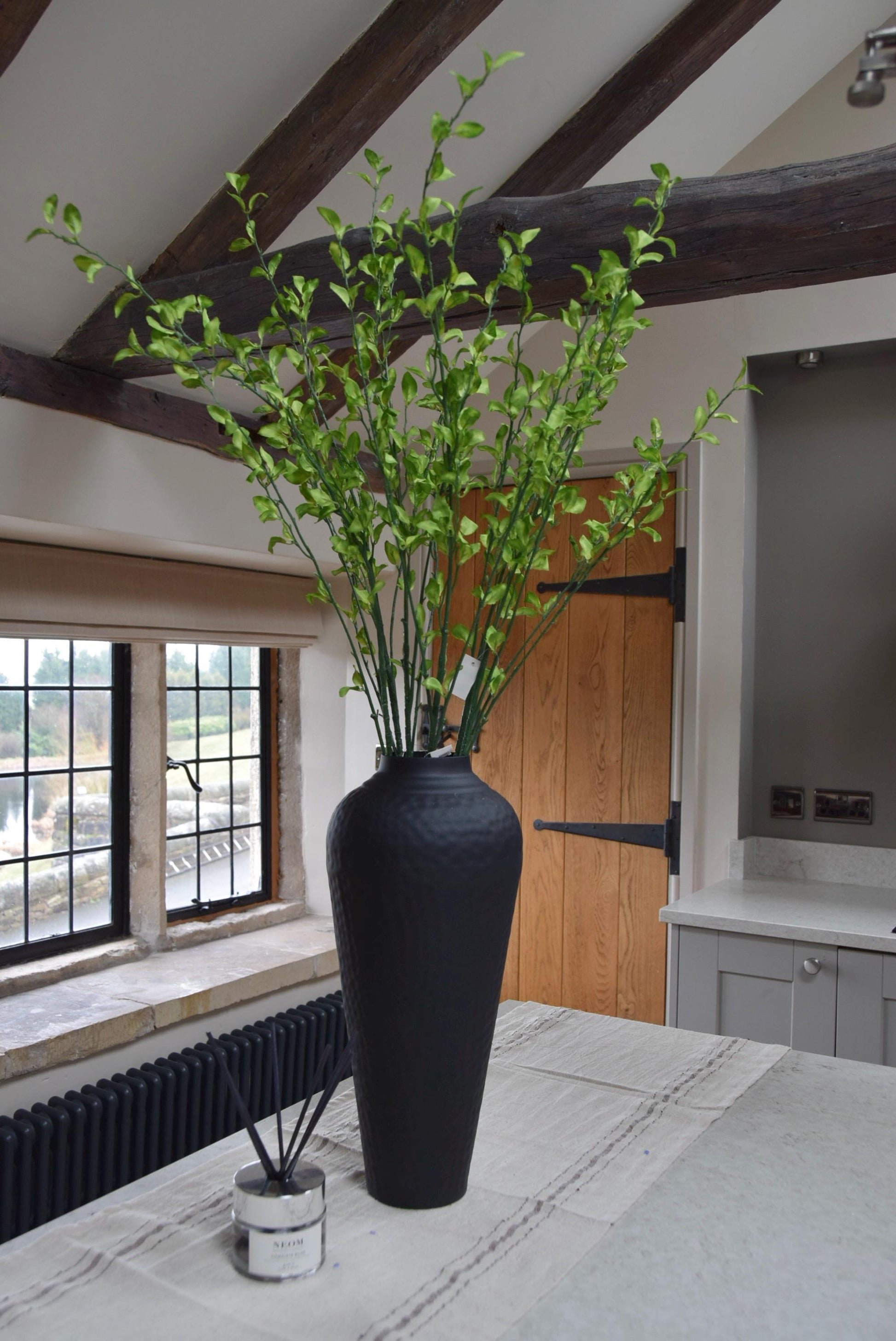 Tall black vase placed on a kitchen countertop with a bunch of green leaf foliage stems. Displayed in a country style kitchen for home decor.