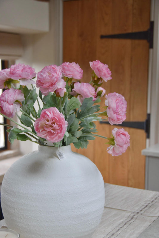 White round vase placed on a kitchen countertop with a bunch of pink ranunculus stems. Displayed in a country style kitchen for home decor.