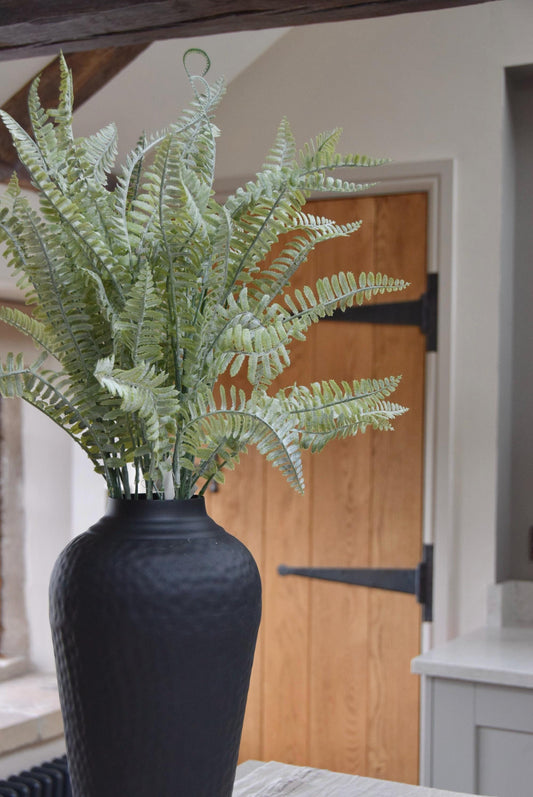 Tall black vase with long dusted fern stems placed on a kitchen countertop in a country style house.