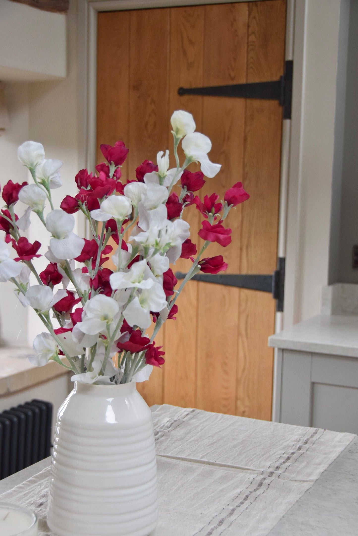 Red and white sweet peas set in a cream vase on a kitchen countertop in a country style kitchen for home decor.