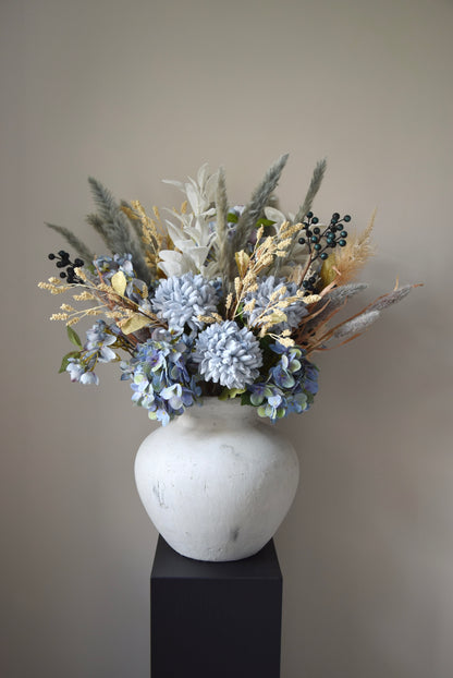 White vase placed on a black pedestal with a bouquet of mixed neutral and grey dried flowers including hydrangeas, pampas grass and lambs ear stems. Styled against a neutral beige wall for home decor.