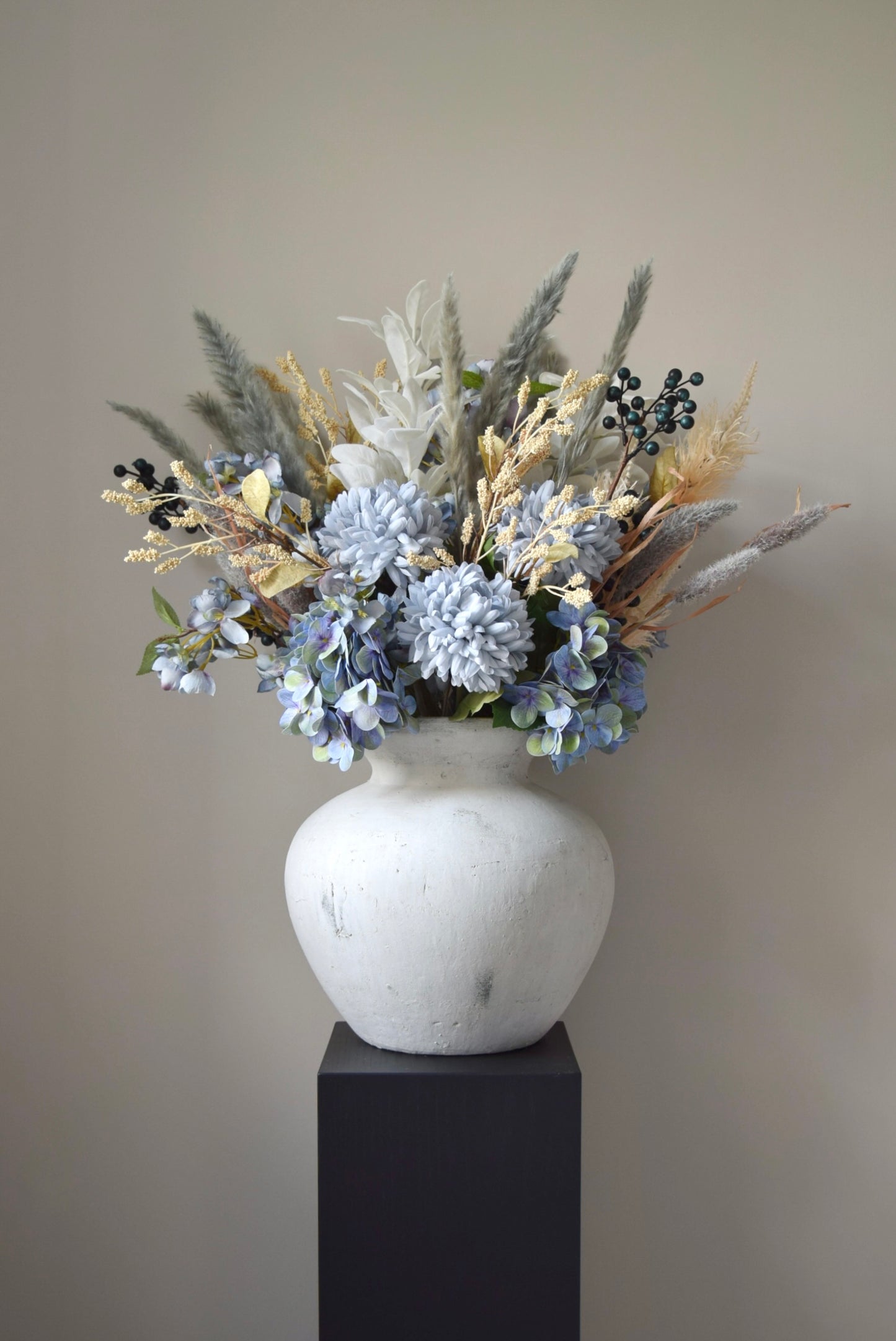 White vase placed on a black pedestal with a bouquet of mixed neutral and grey dried flowers including hydrangeas, pampas grass and lambs ear stems. Styled against a neutral beige wall for home decor.