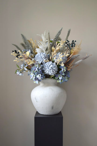 White vase placed on a black pedestal with a bouquet of mixed neutral and grey dried flowers including hydrangeas, pampas grass and lambs ear stems. Styled against a neutral beige wall for home decor.