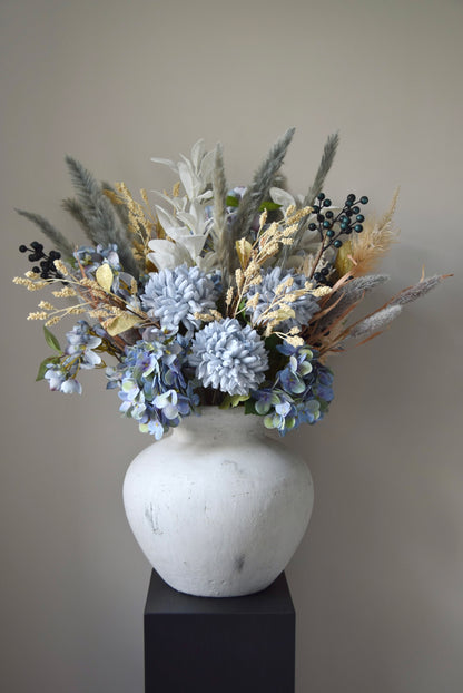 White vase placed on a black pedestal with a bouquet of mixed neutral and grey dried flowers including hydrangeas, pampas grass and lambs ear stems. Styled against a neutral beige wall for home decor.