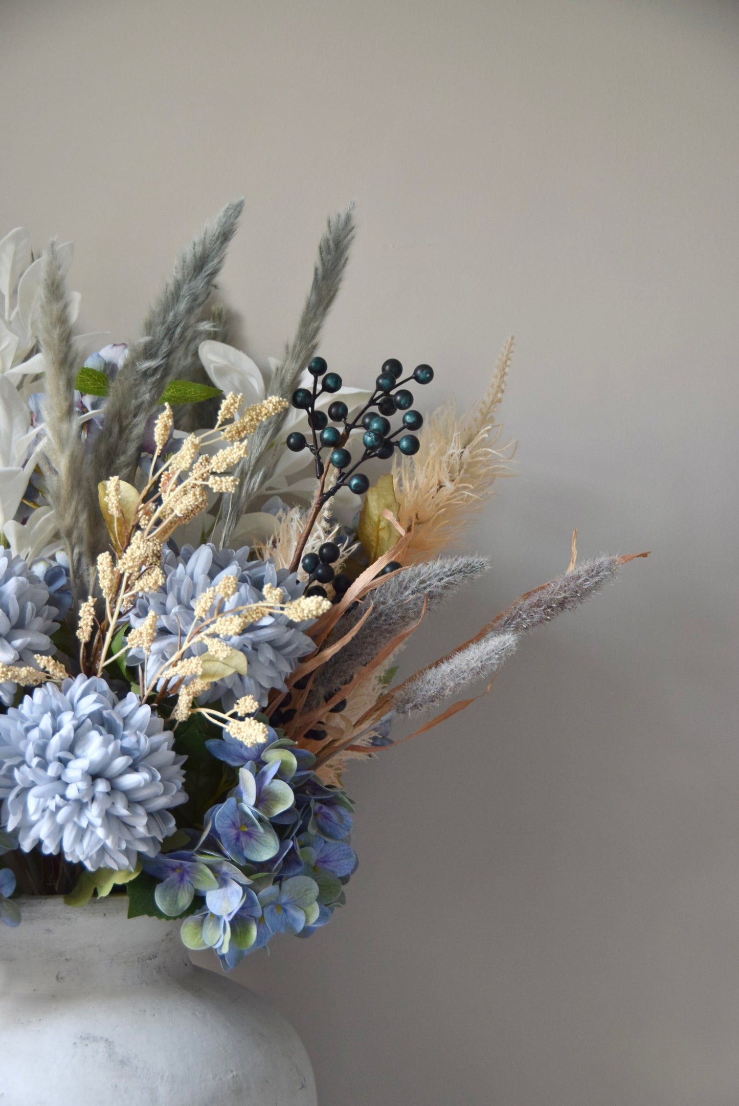 White vase placed on a black pedestal with a bouquet of mixed neutral and grey dried flowers including hydrangeas, pampas grass and lambs ear stems. Styled against a neutral beige wall for home decor.