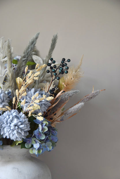 White vase placed on a black pedestal with a bouquet of mixed neutral and grey dried flowers including hydrangeas, pampas grass and lambs ear stems. Styled against a neutral beige wall for home decor.