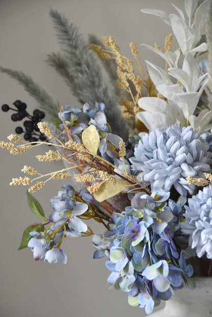 White vase placed on a black pedestal with a bouquet of mixed neutral and grey dried flowers including hydrangeas, pampas grass and lambs ear stems. Styled against a neutral beige wall for home decor.