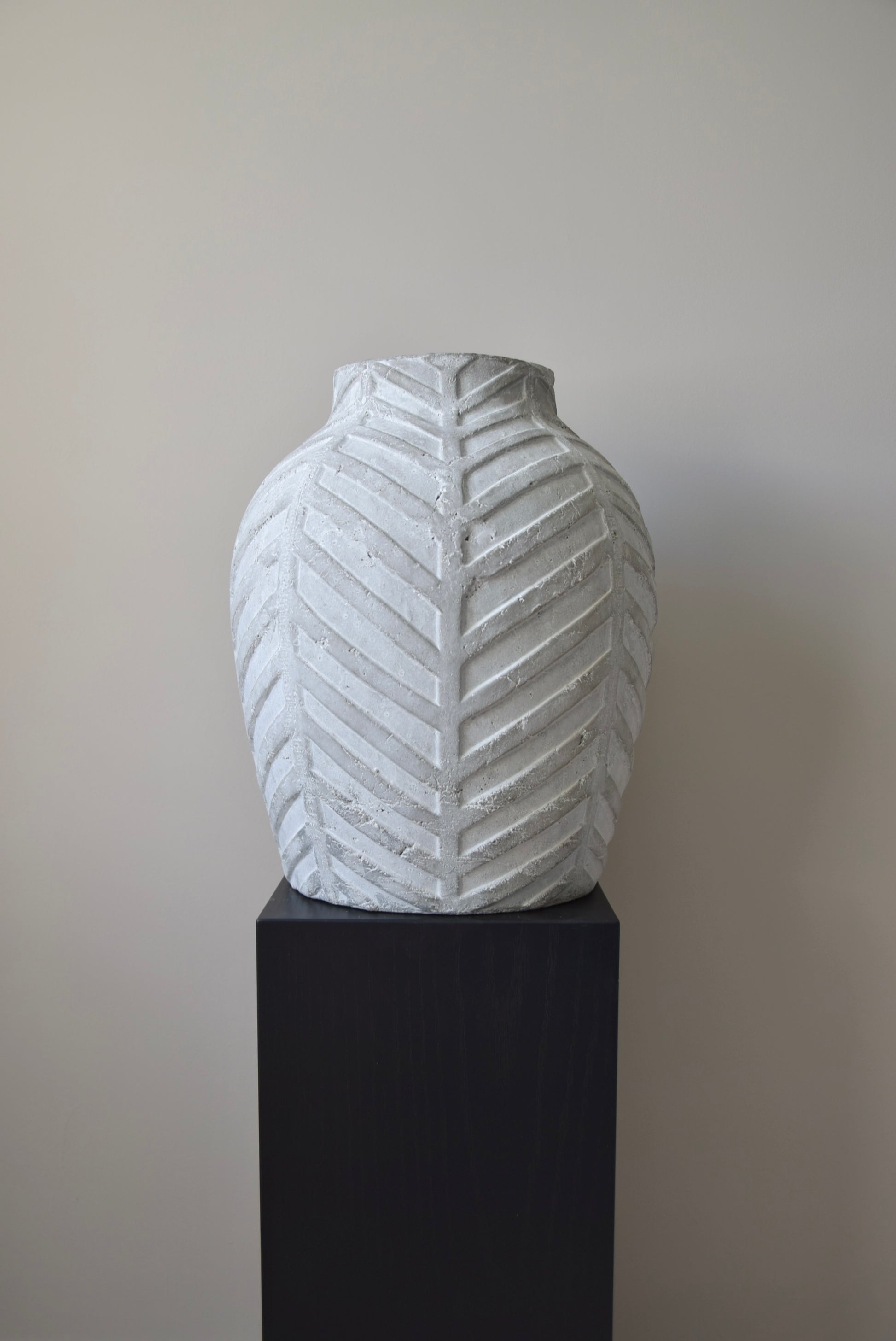 Grey chevron stone ceramic vase displayed on a black pedestal against a beige wall.