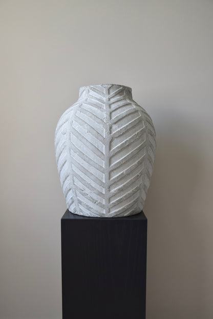 Grey chevron stone ceramic vase displayed on a black pedestal against a beige wall.
