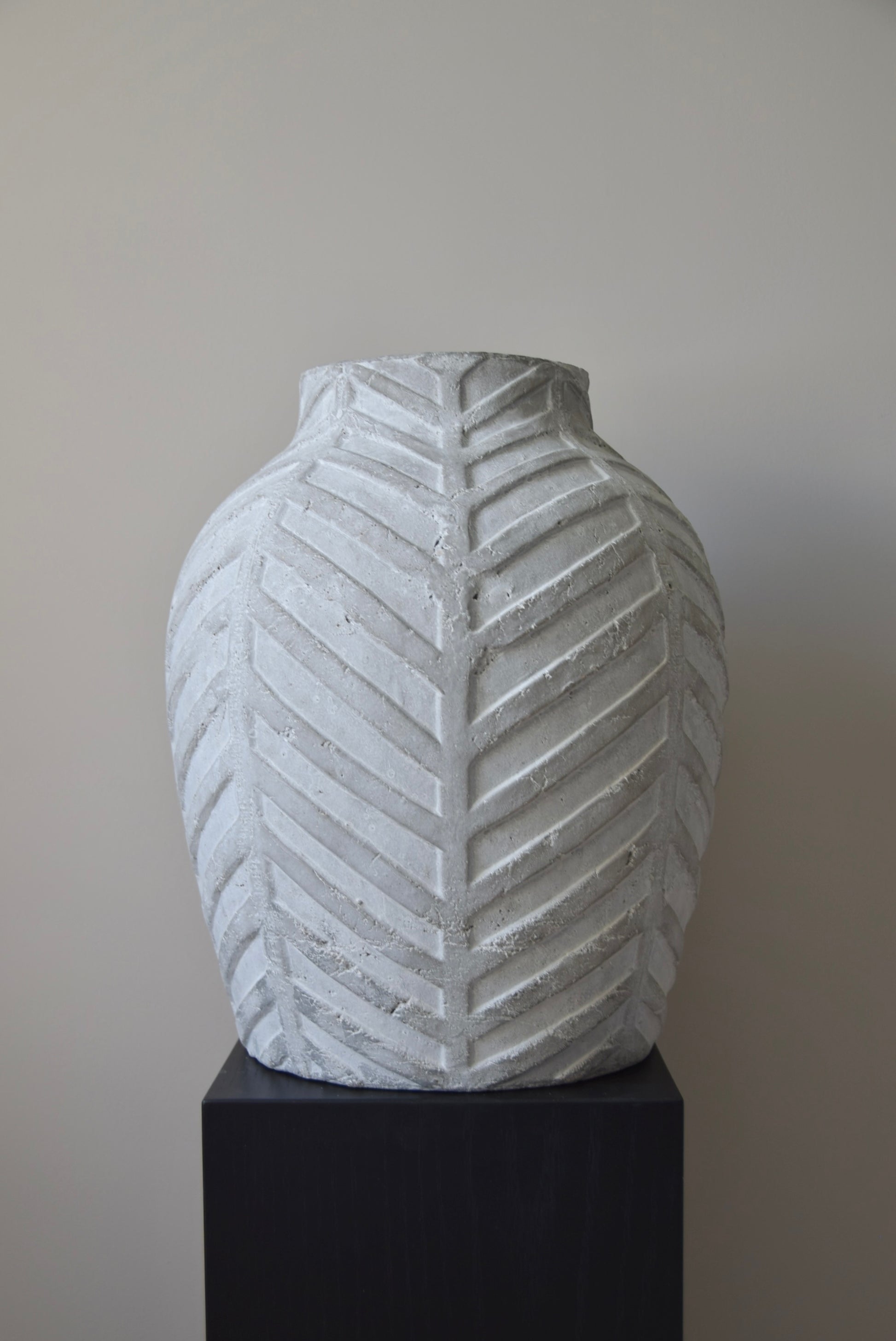 Grey chevron stone ceramic vase displayed on a black pedestal against a beige wall.