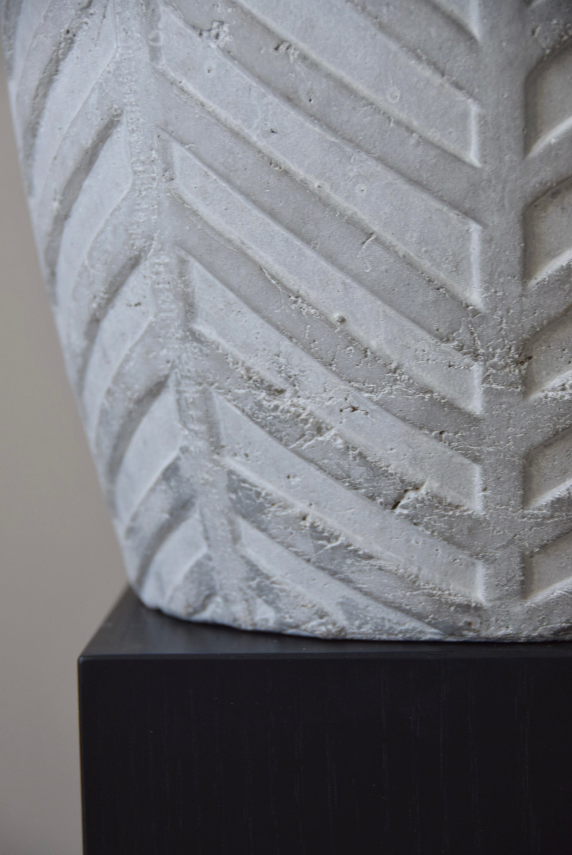 Grey chevron stone ceramic vase displayed on a black pedestal against a beige wall.