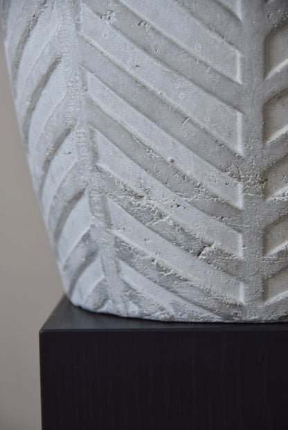 Grey chevron stone ceramic vase displayed on a black pedestal against a beige wall.