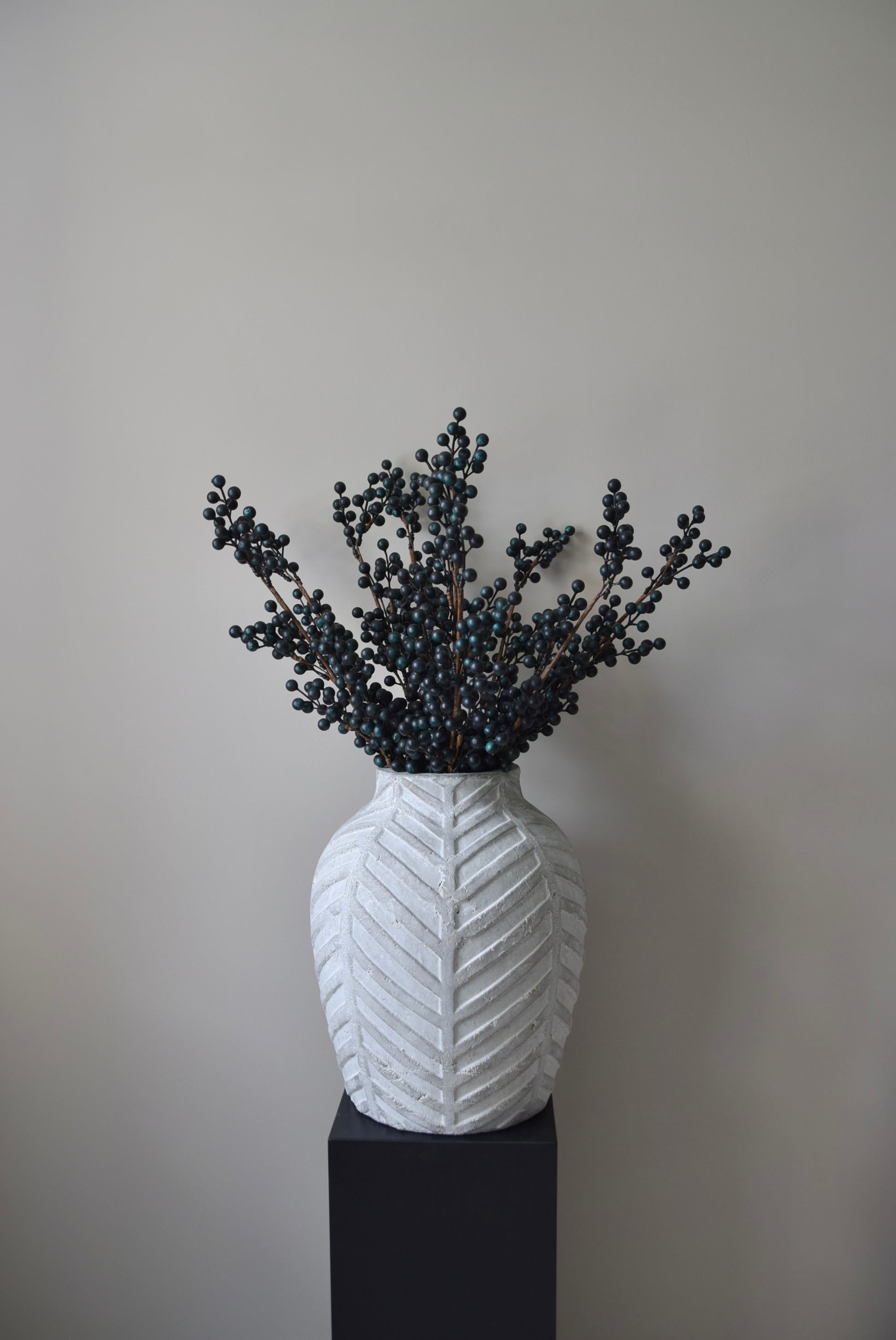 Stone chevron style vase placed on a black pedestal displaying some blueberry stems.