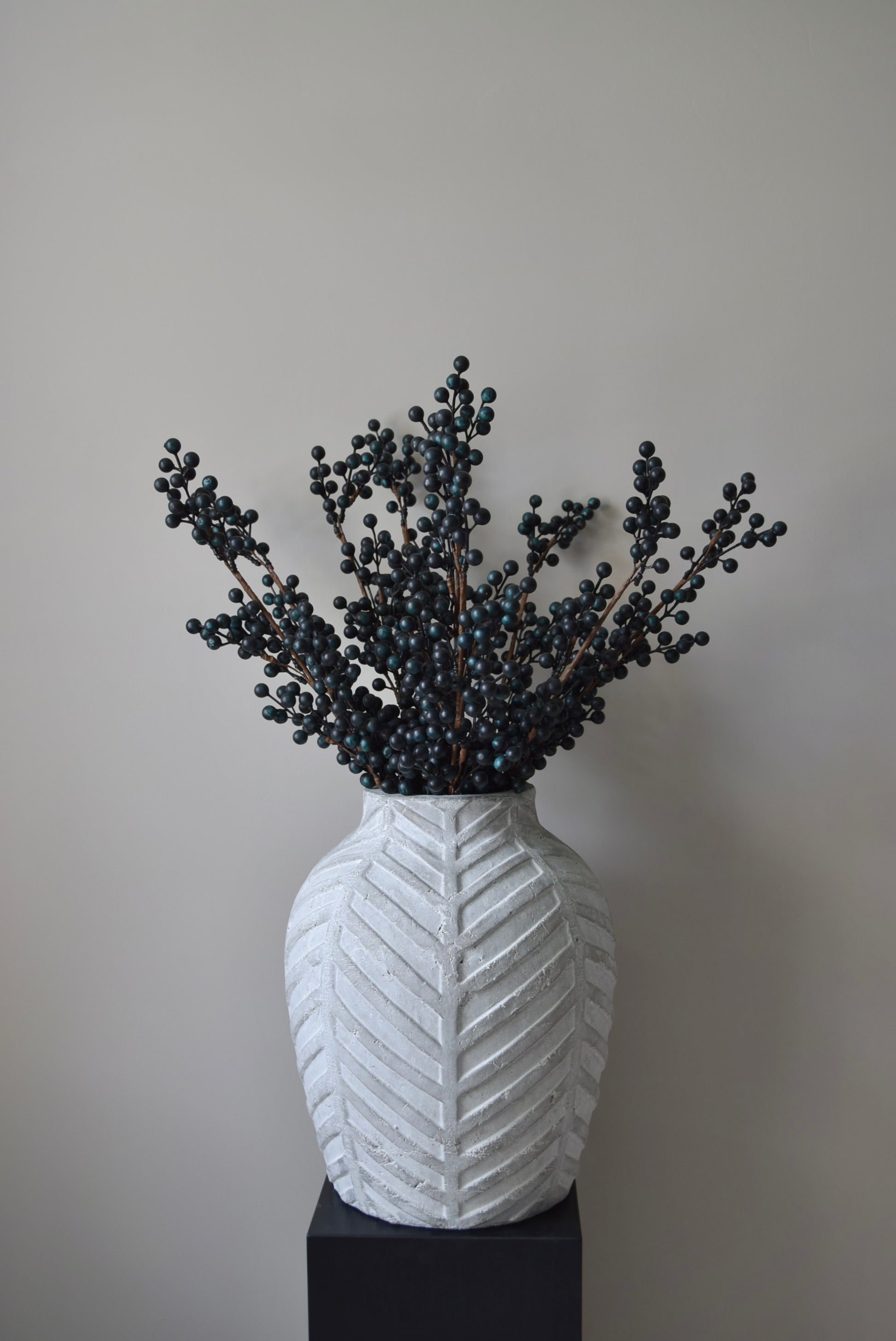 Stone chevron style vase placed on a black pedestal displaying some blueberry stems.