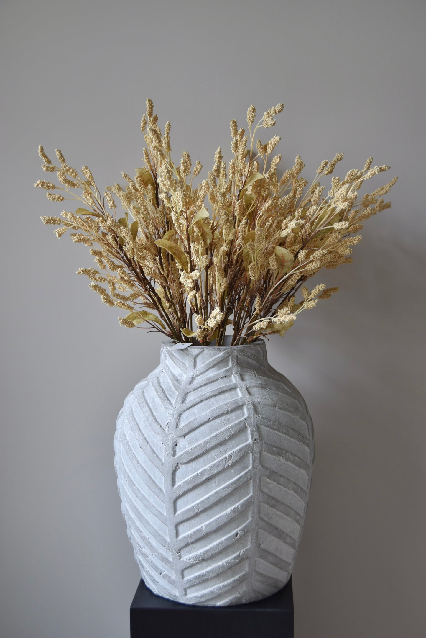 Dried-look grain sorghum spray displayed in a stone chevron style vase placed on a black pedestal.