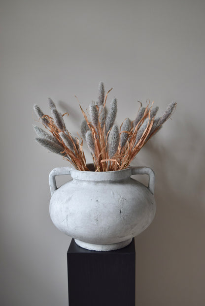 Grey dried look bunny tail reed stems displayed in a greek pelike stone pot placed on a black pedestal.