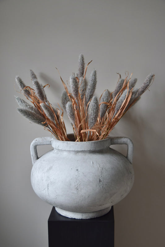 Grey dried look bunny tail reed stems displayed in a greek pelike stone pot placed on a black pedestal.
