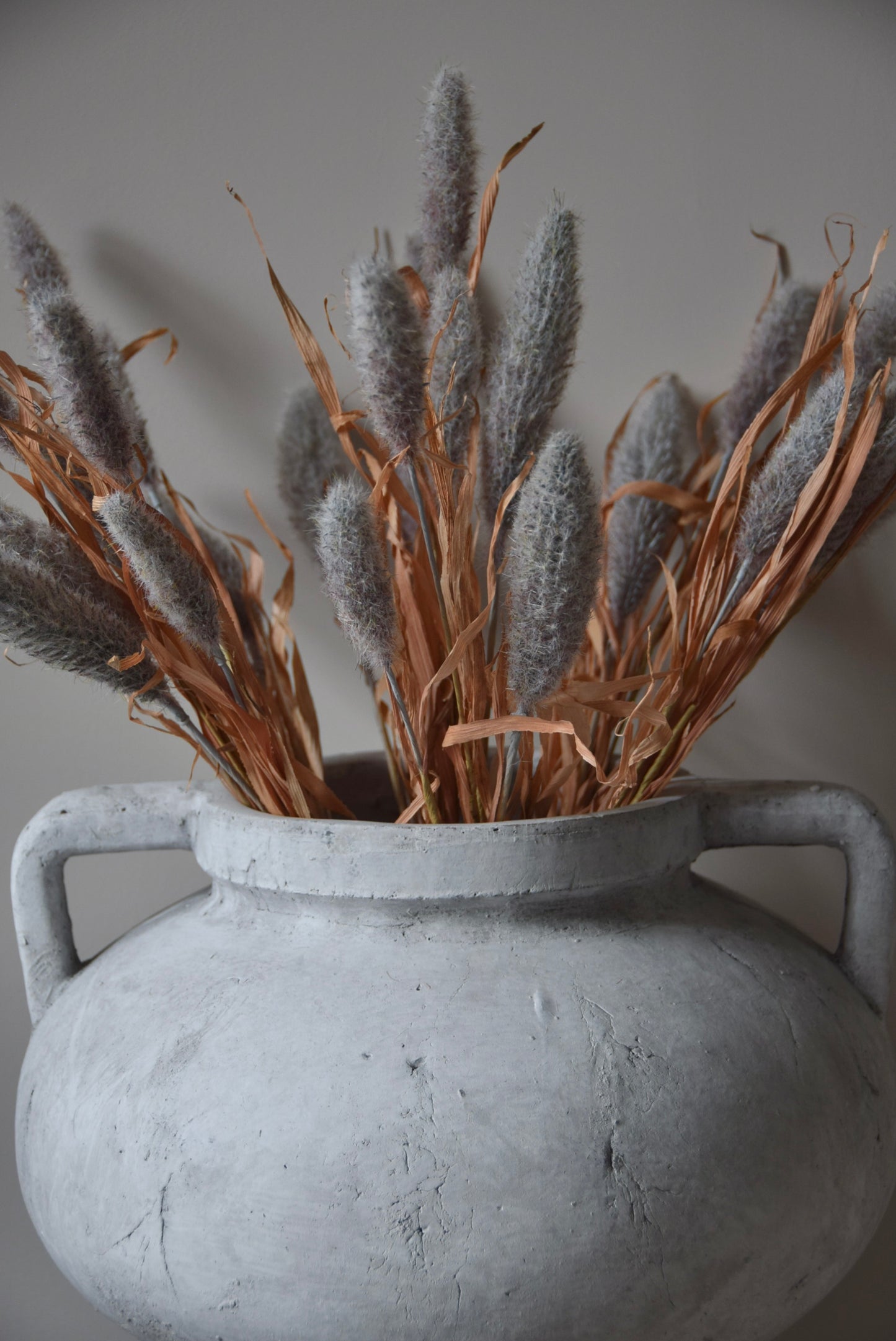 Grey dried look bunny tail reed stems displayed in a greek pelike stone pot placed on a black pedestal.