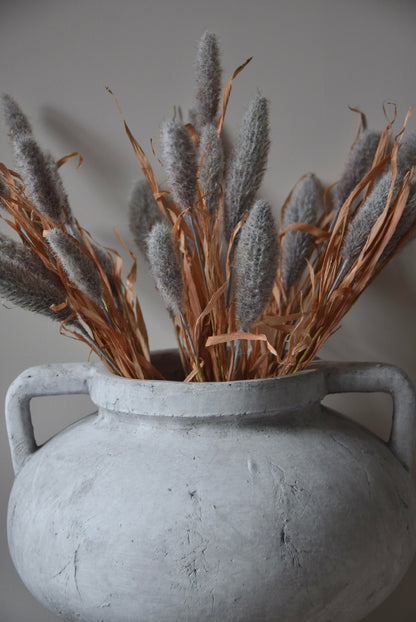 Grey dried look bunny tail reed stems displayed in a greek pelike stone pot placed on a black pedestal.