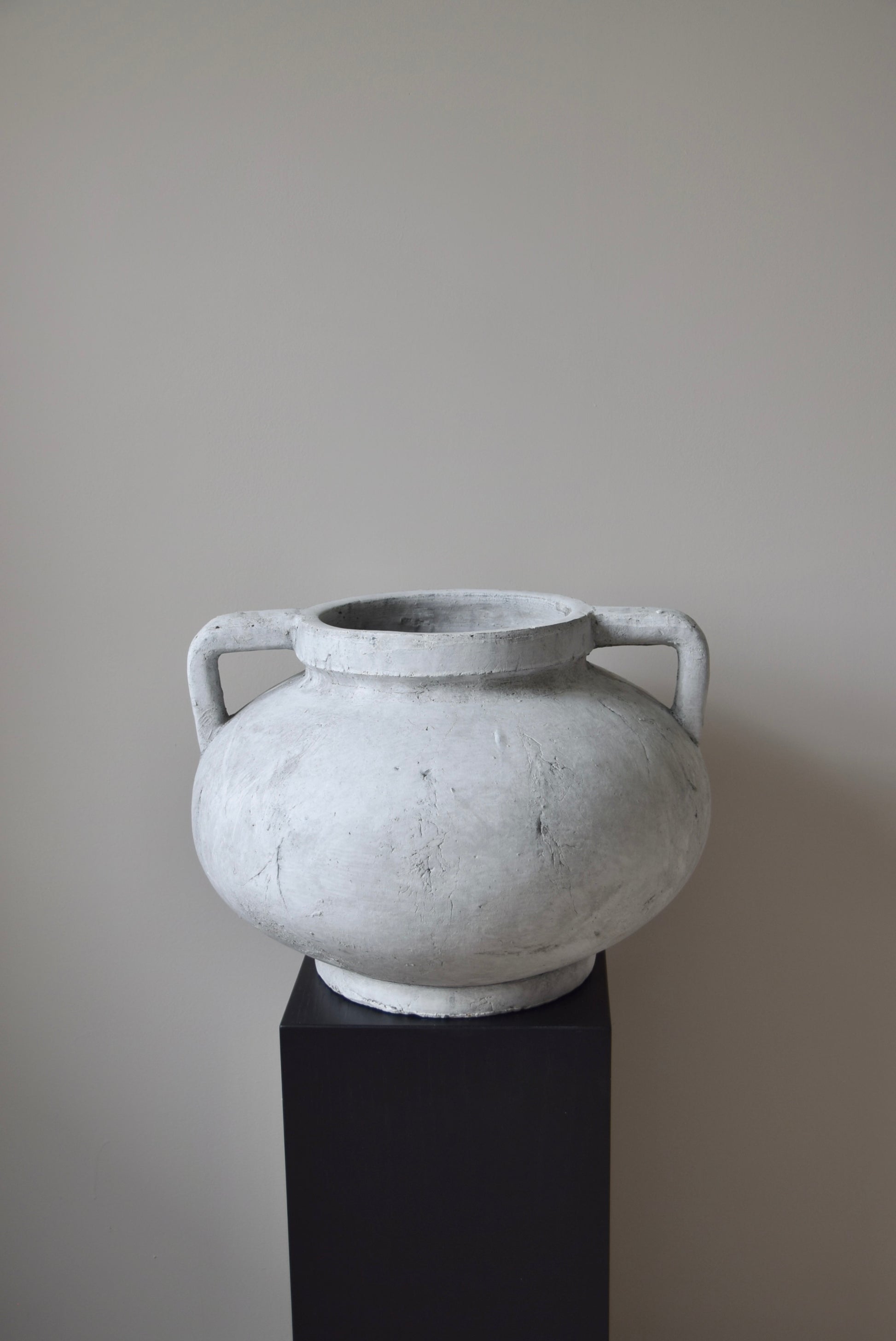 Grey stone Greek pelike vase with two handles displayed on a black pedestal.