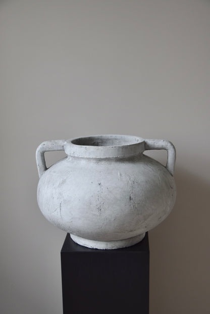 Grey stone Greek pelike vase with two handles displayed on a black pedestal.