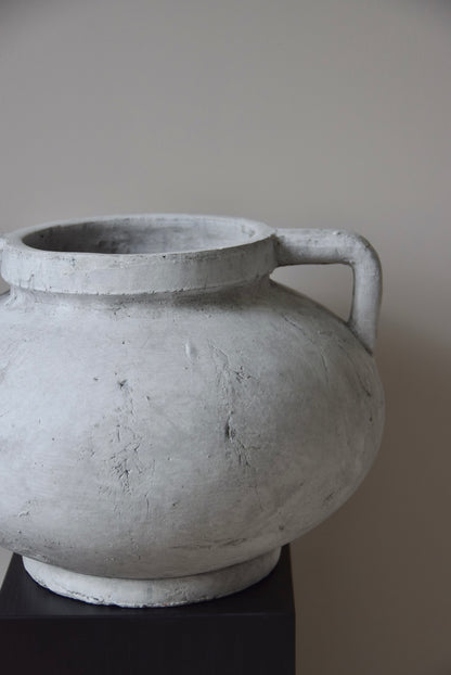 Grey stone Greek pelike vase with two handles displayed on a black pedestal.