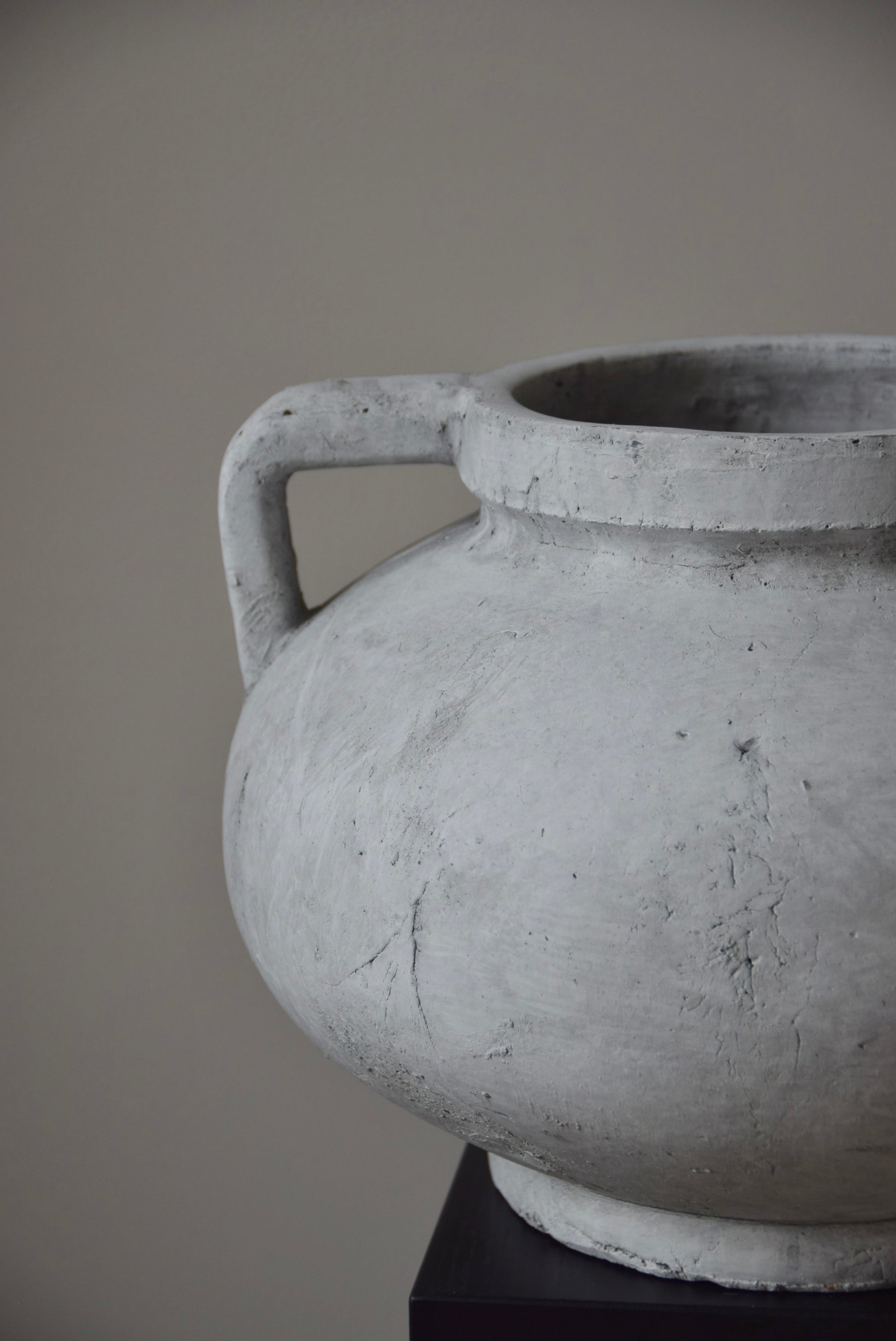Grey stone Greek pelike vase with two handles displayed on a black pedestal.