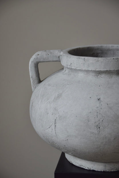 Grey stone Greek pelike vase with two handles displayed on a black pedestal.