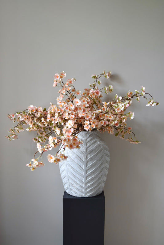 Stone chevron style vase placed on a black pedestal with a bunch of drooping pink plum blossom stems.