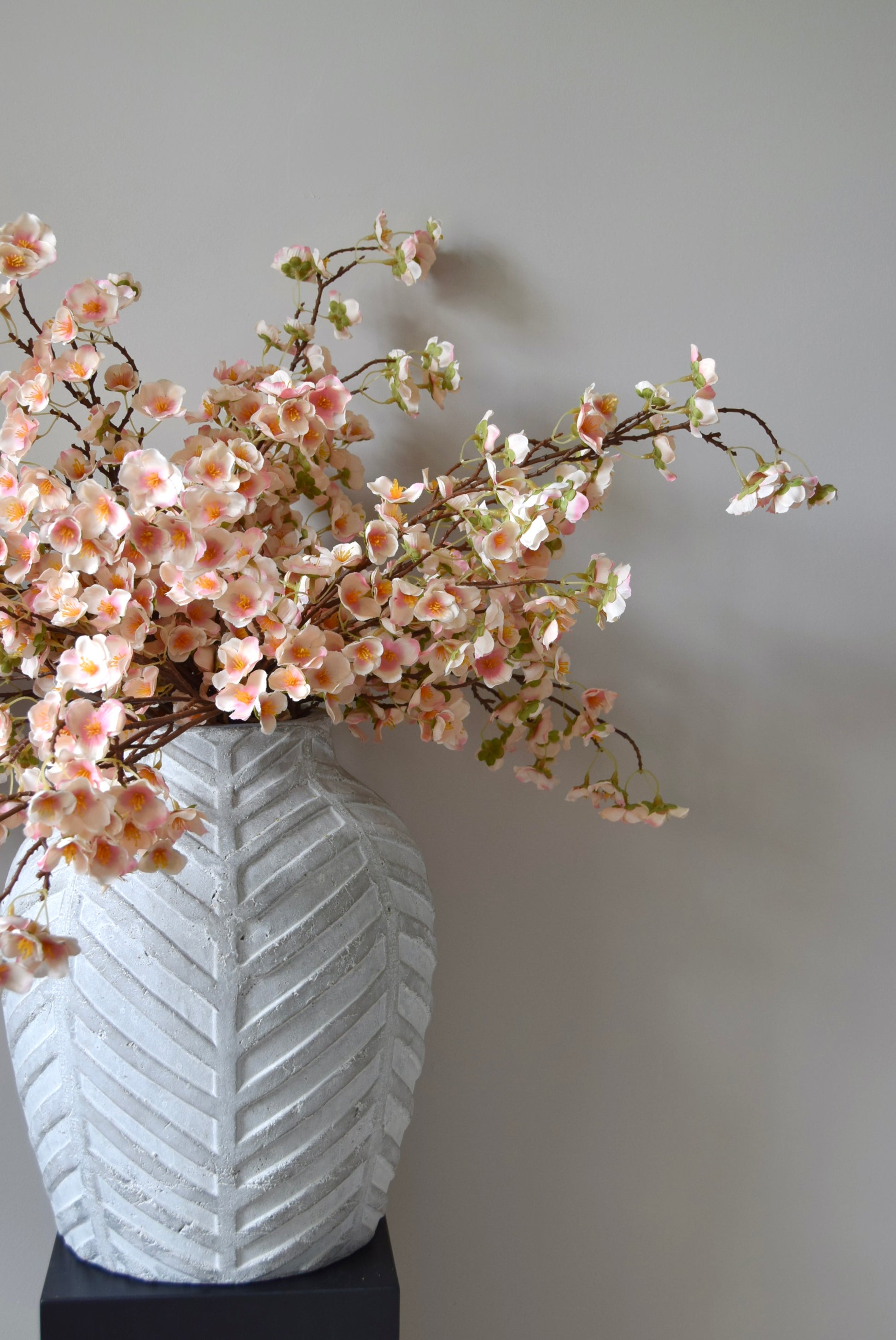 Stone chevron style vase placed on a black pedestal with a bunch of drooping pink plum blossom stems.