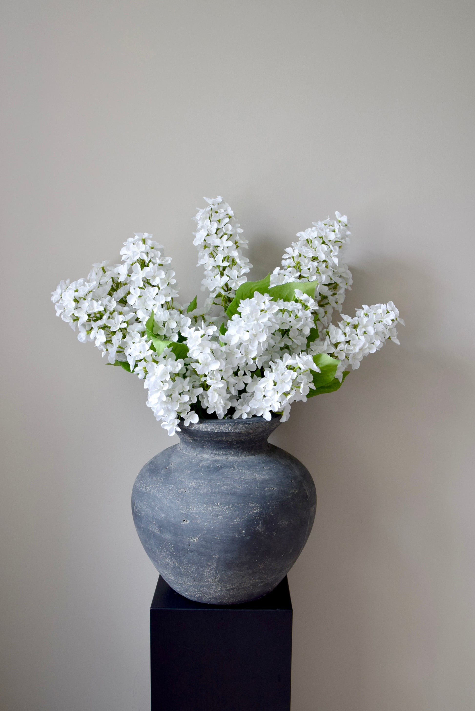 Grey vase with white flowers on a black stand against a plain background