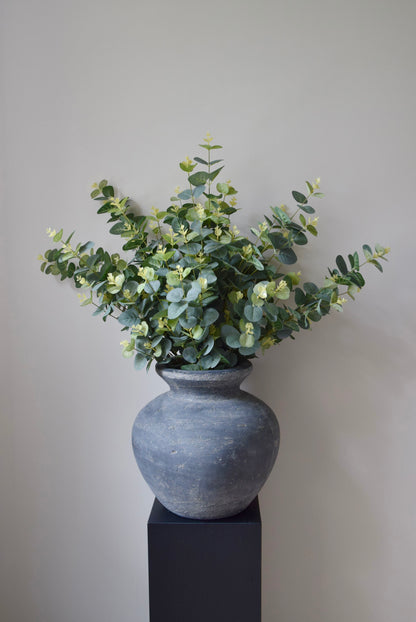 Grey textured vase with greenery on a black pedestal against a plain wall