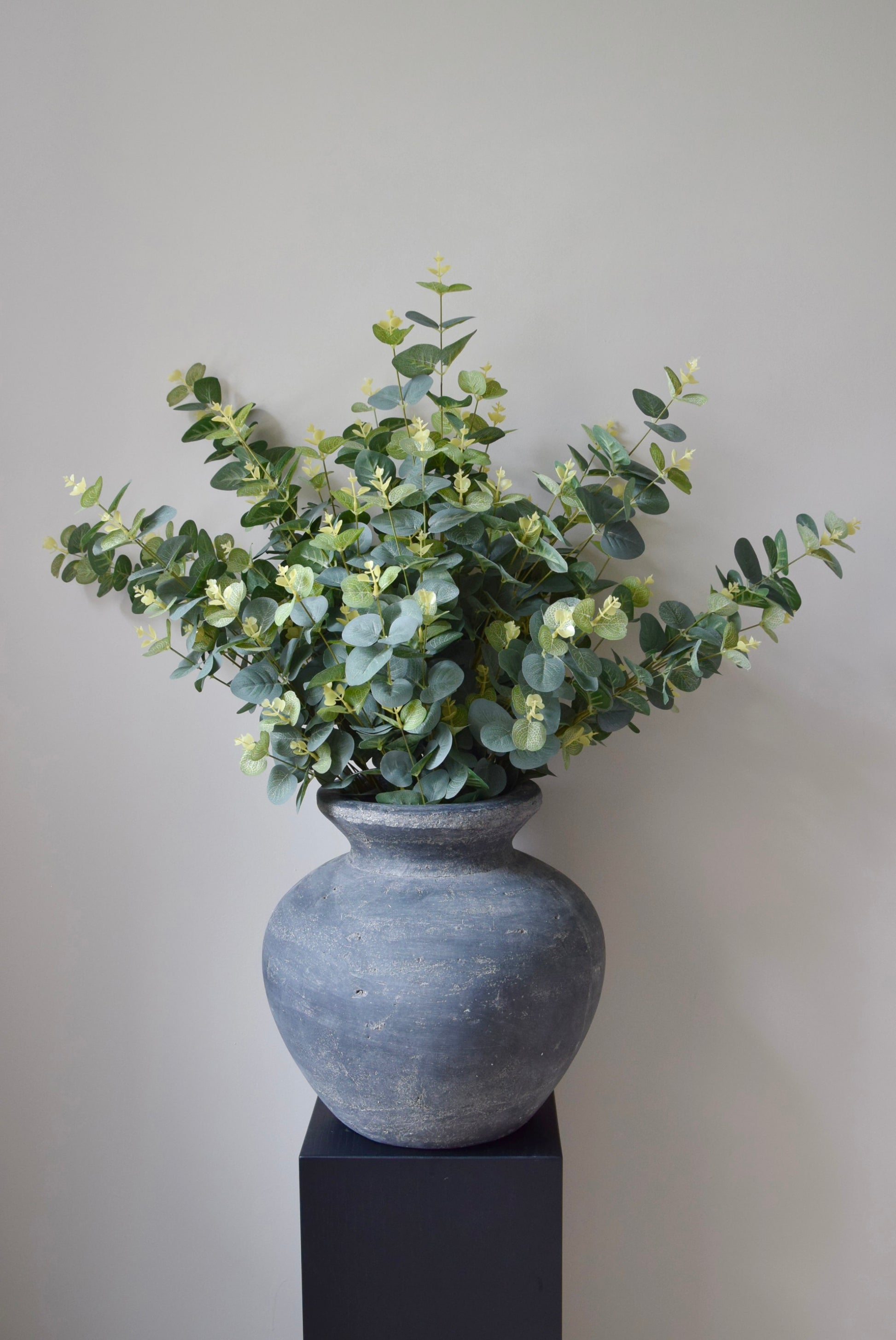 Grey textured vase with greenery on a black stand against a plain background