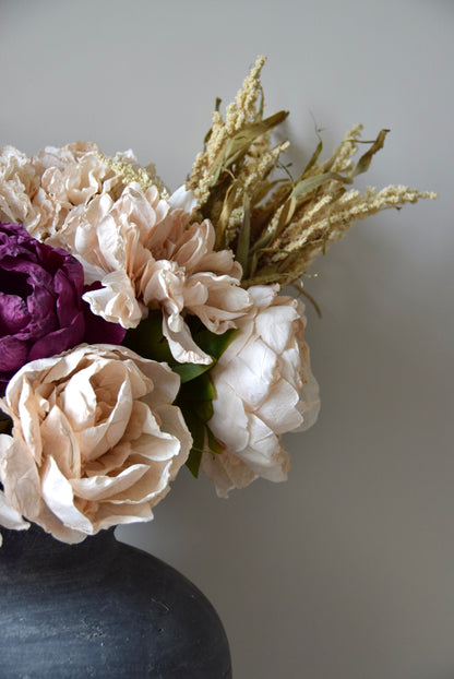 Floral arrangement with beige, purple, and gold flowers in a dark vase on a light gray background