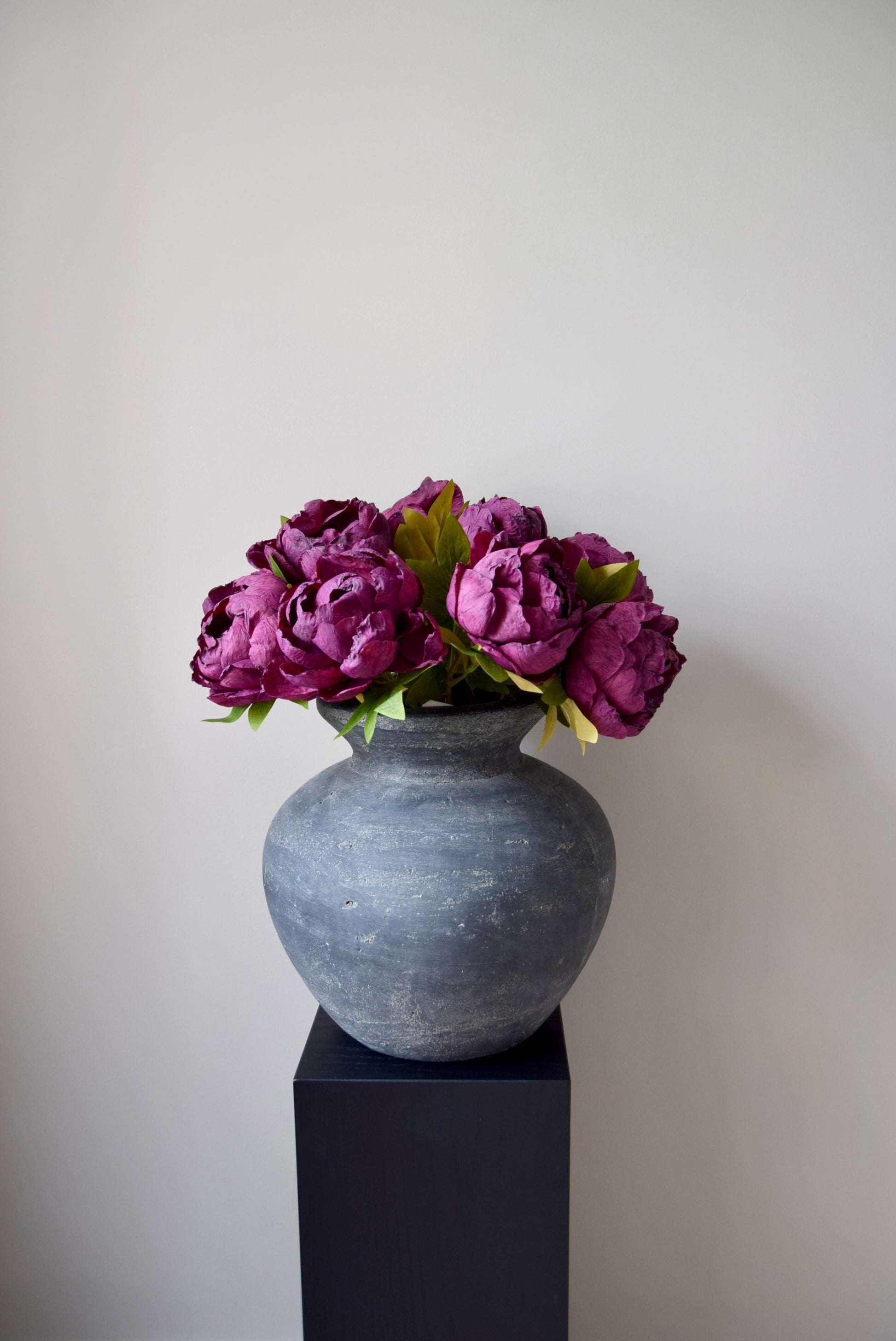 Grey vase with purple flowers on a black pedestal against a plain background