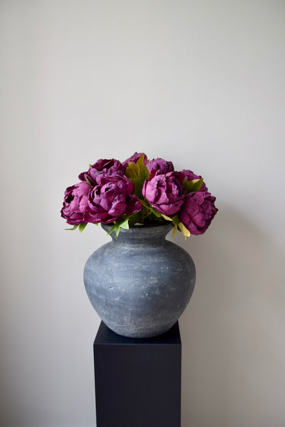 Grey vase with purple flowers on a black pedestal against a plain background
