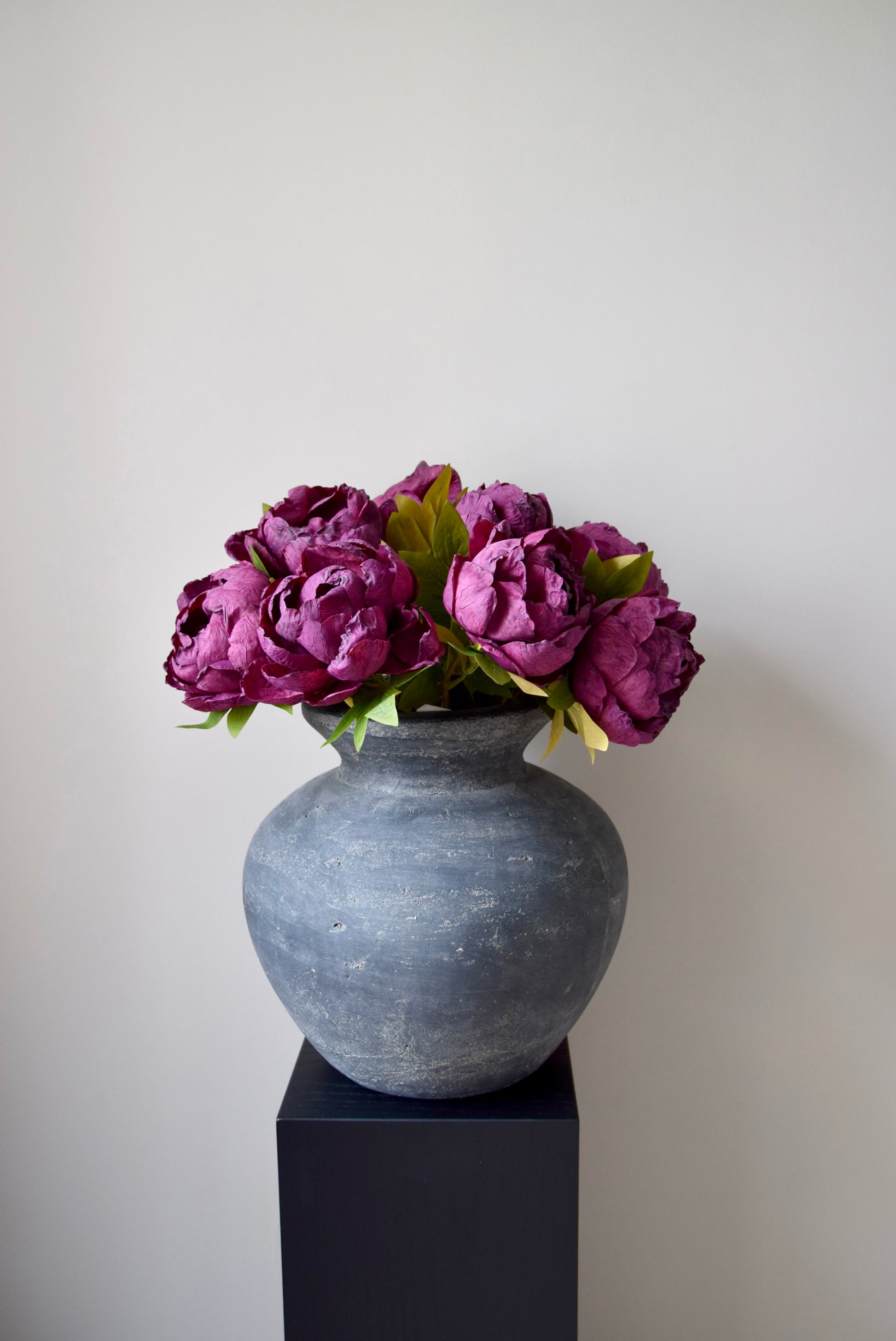 Bunch of burgundy peonies in a grey vase on a black pedestal.