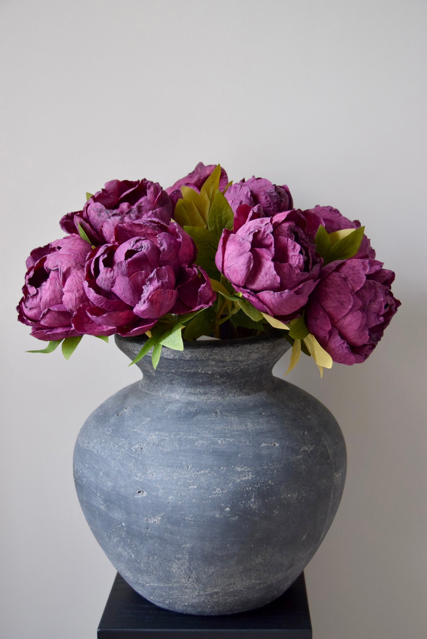 Bunch of burgundy peonies in a grey vase on a black pedestal.