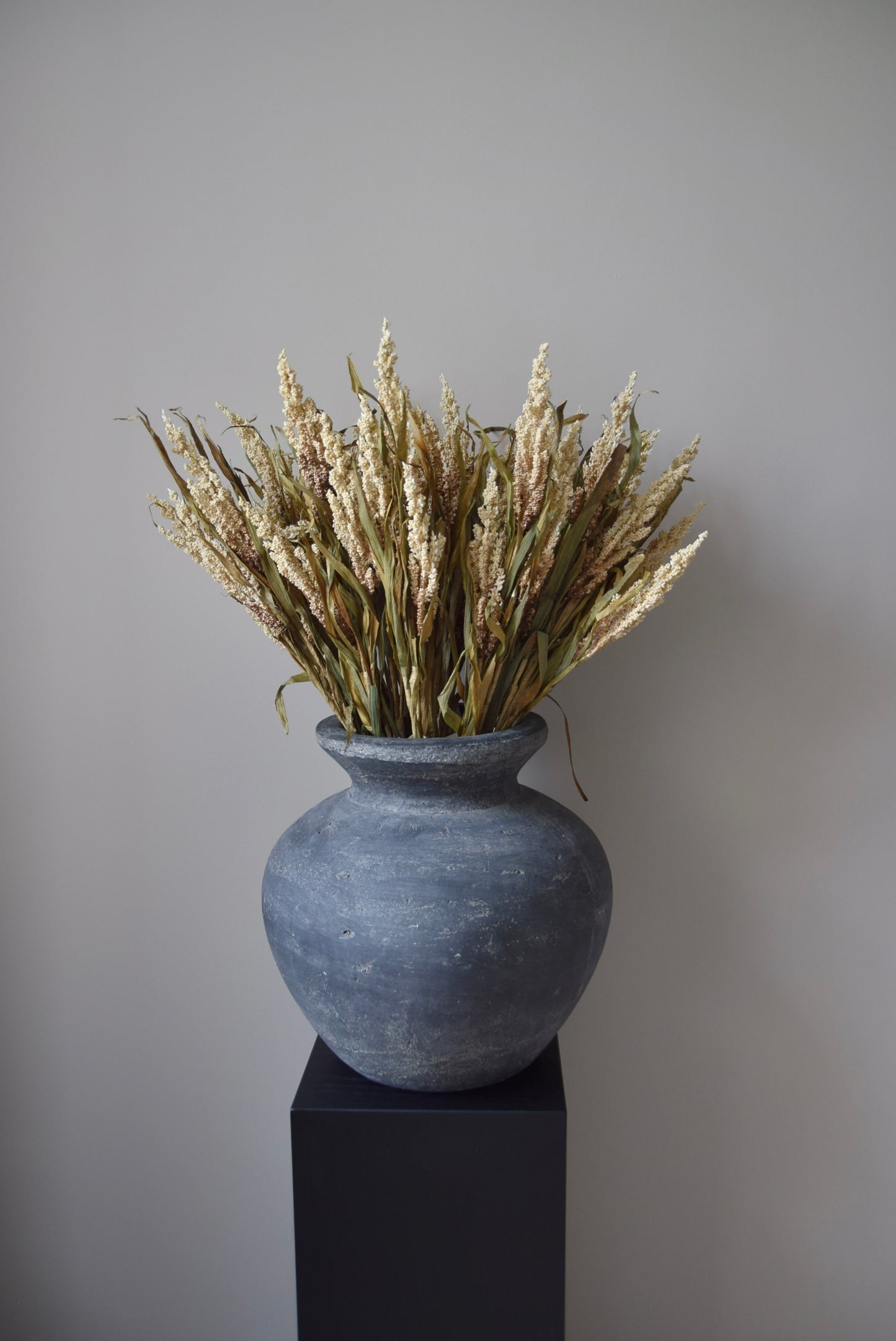 Grey vase with dried plants on a black stand against a plain wall