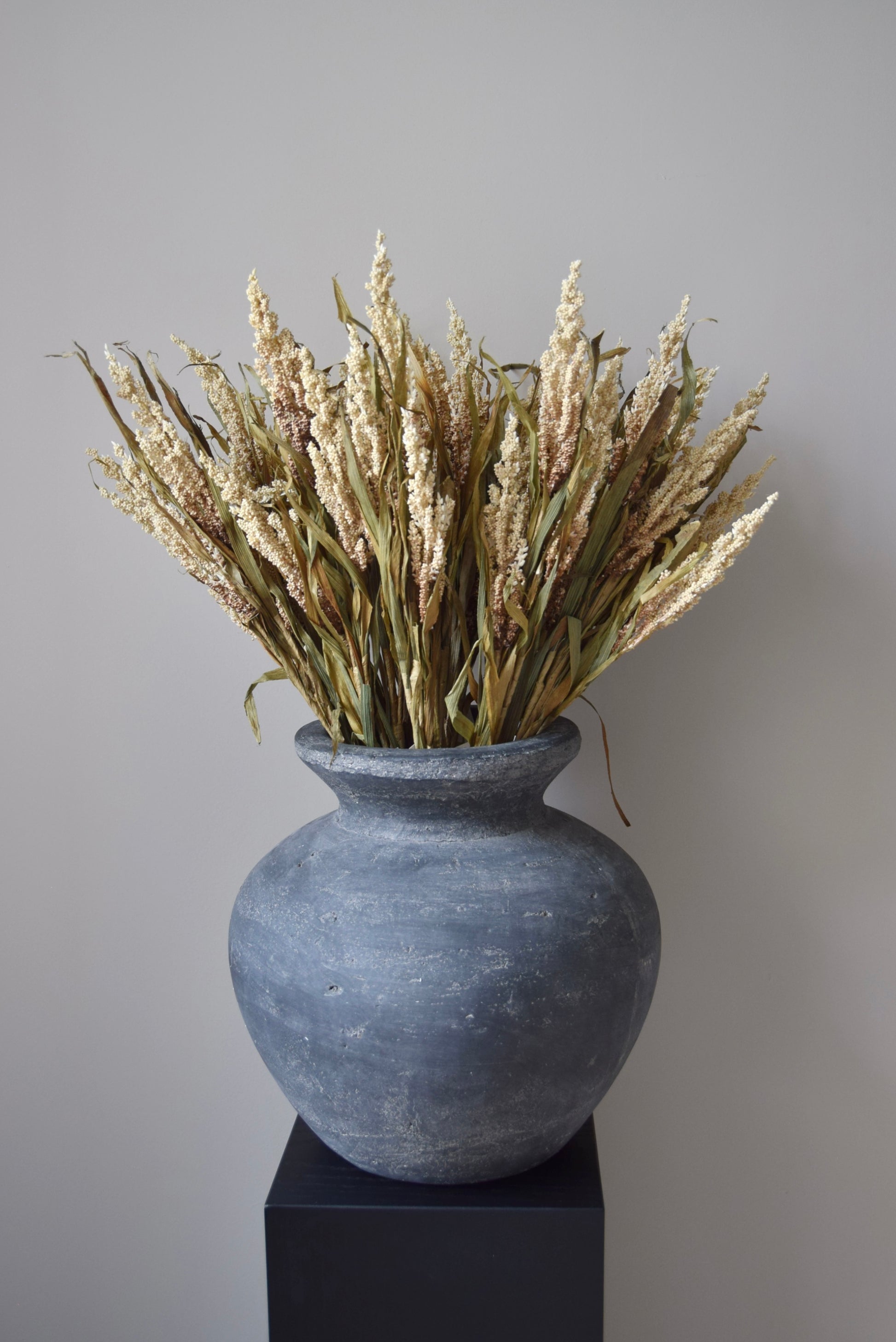 Grey vase with dried plants on a black stand against a neutral background