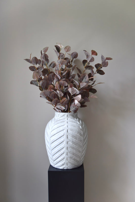 White textured vase with dried plants on a black stand against a plain background